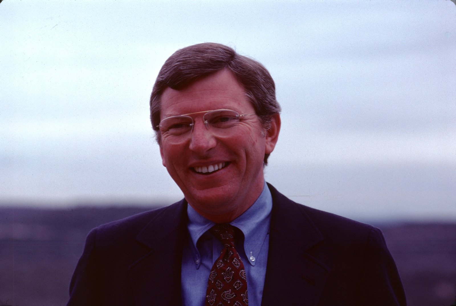 A young Mark White smiling outdoors in a navy suit, 1970s.