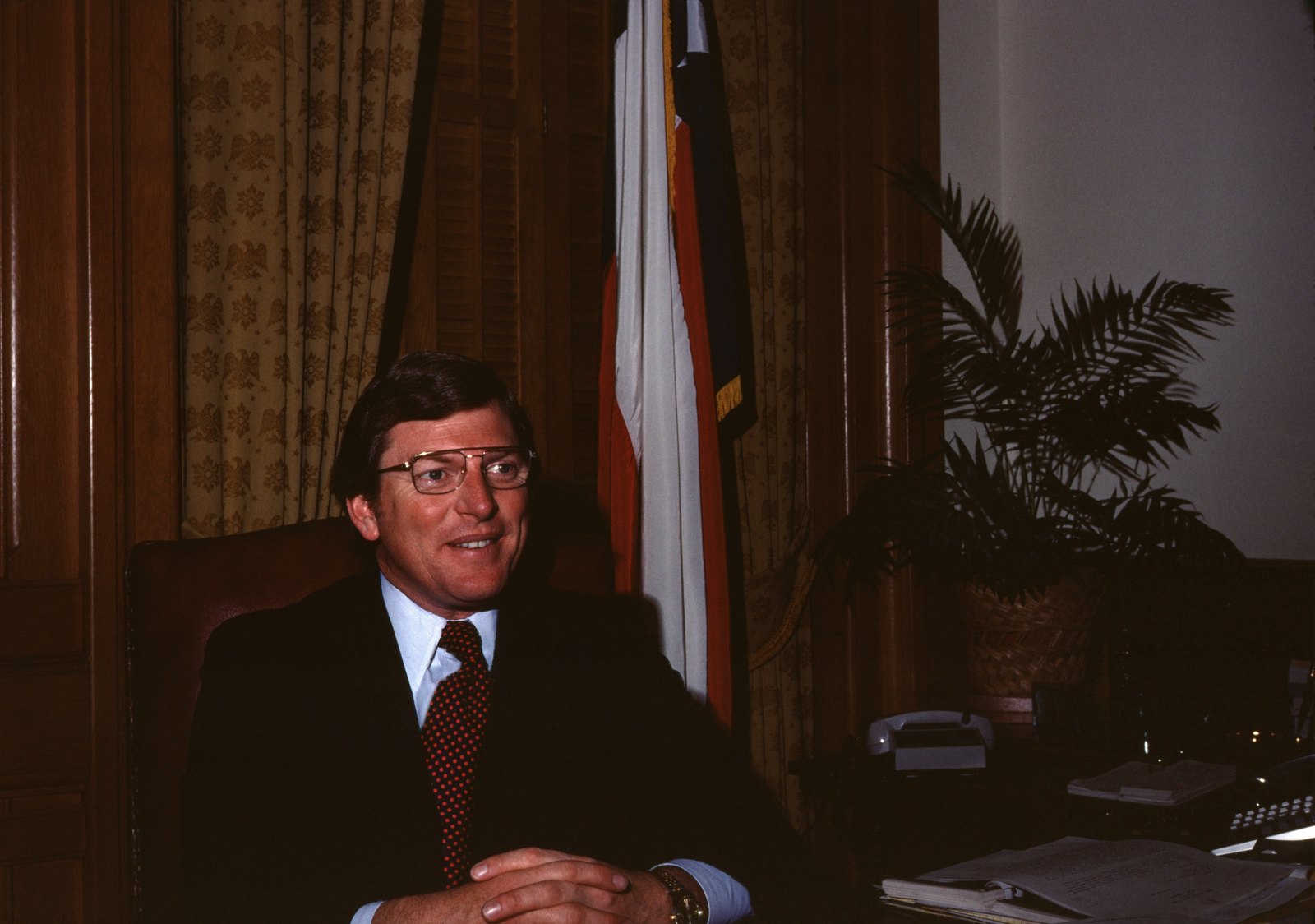 Mark White seated at his desk beside the Texas flag.