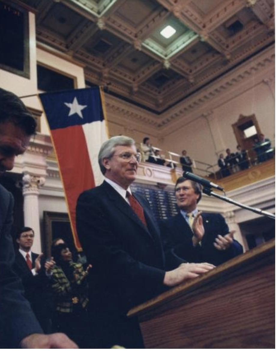 Governor Mark White speaking at the podium of the Texas House of Representatives, with the Texas flag behind him.